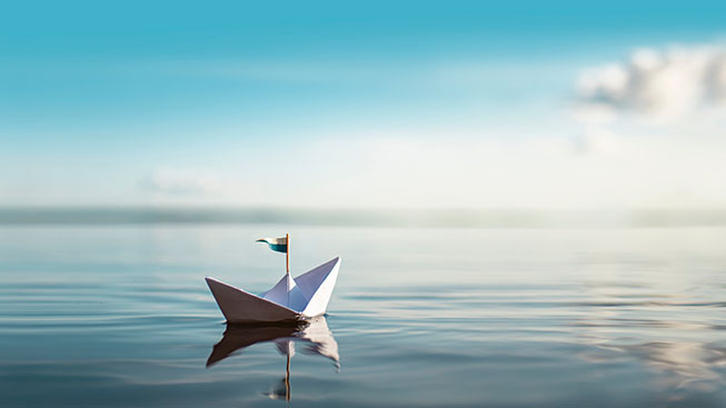 A paper boat with a small flag floats on a body of water. Above it a blue sky with white clouds.